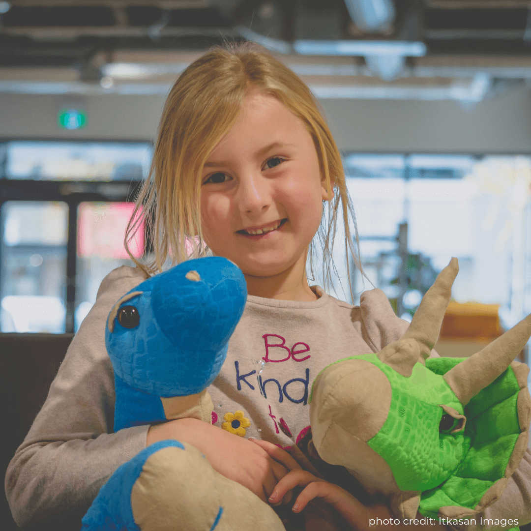 Image of a young, smiling girl with dinosaur stuffed animals. Book your next birthday party with Dino Lab Inc!
